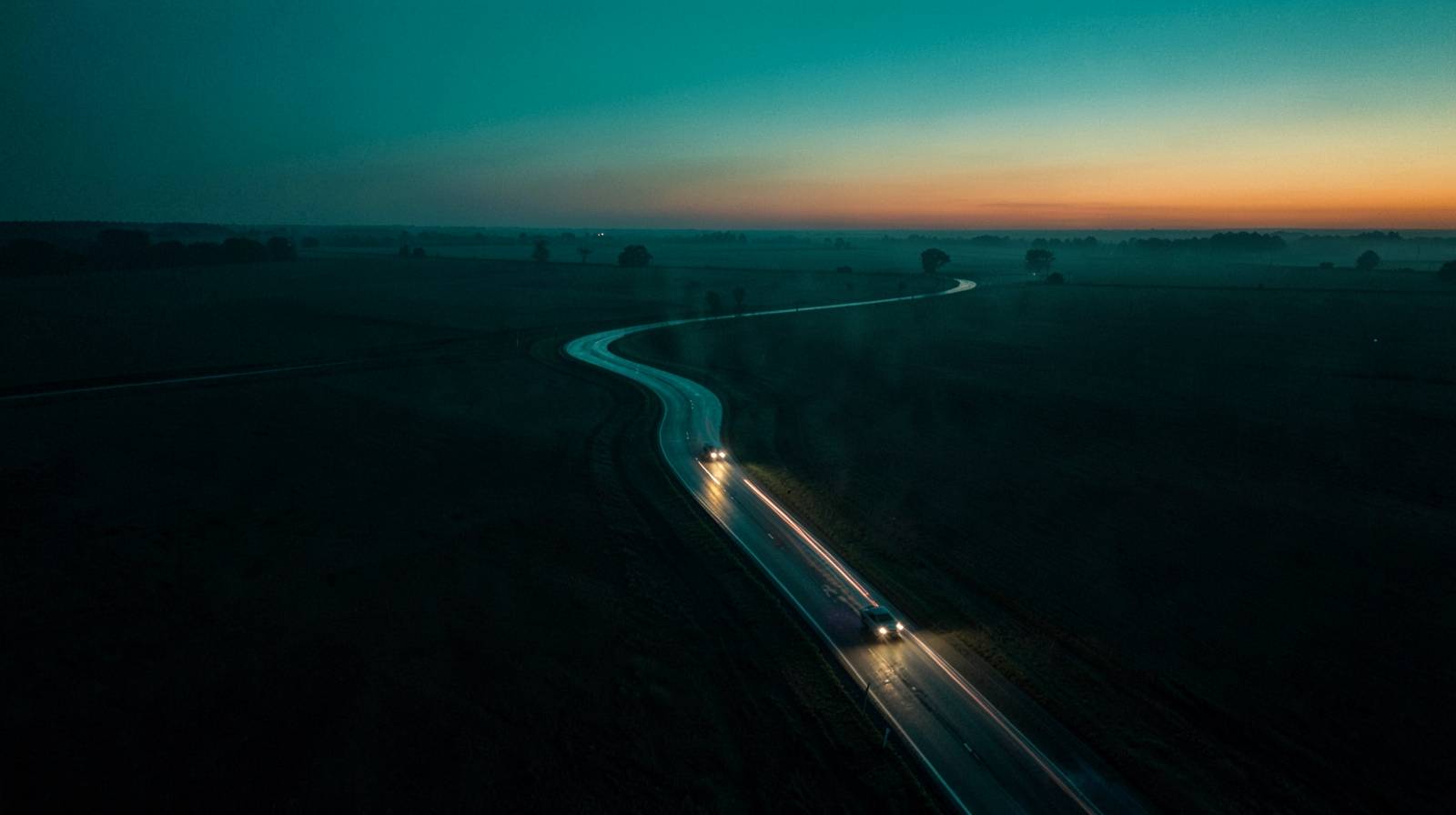 A single road winding through dark farmland at dusk, headlights following the curve